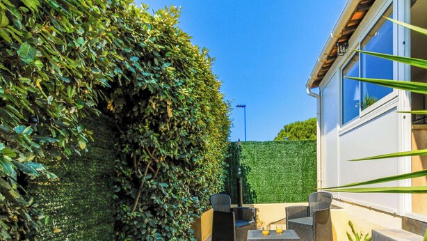 Une maison avec une haie bien entretenue, un espace extérieur aménagé pour s’asseoir et une vue dégagée sur le ciel.