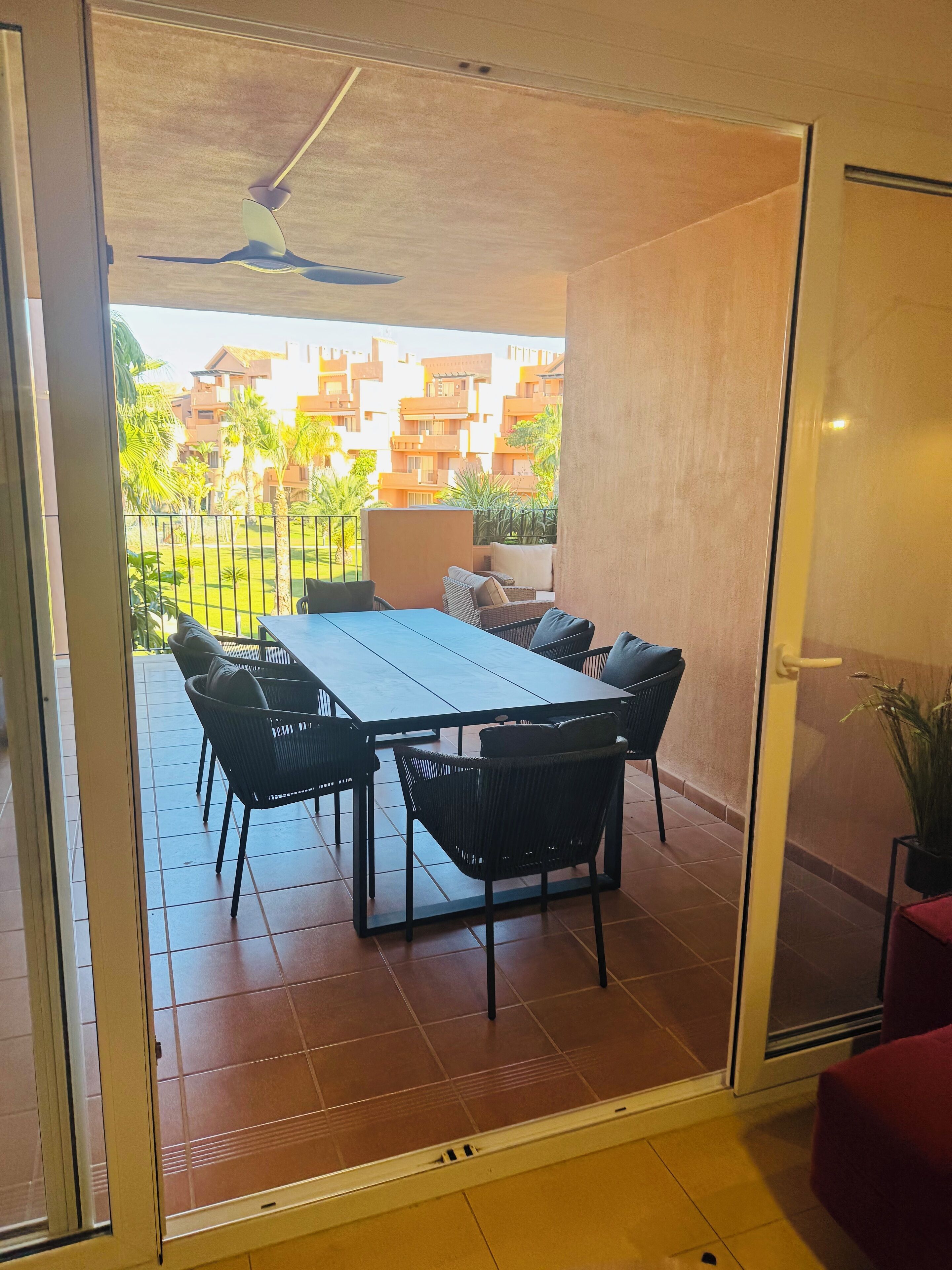 A dining area with a table and chairs, a ceiling fan, and a view of a residential complex with palm trees.