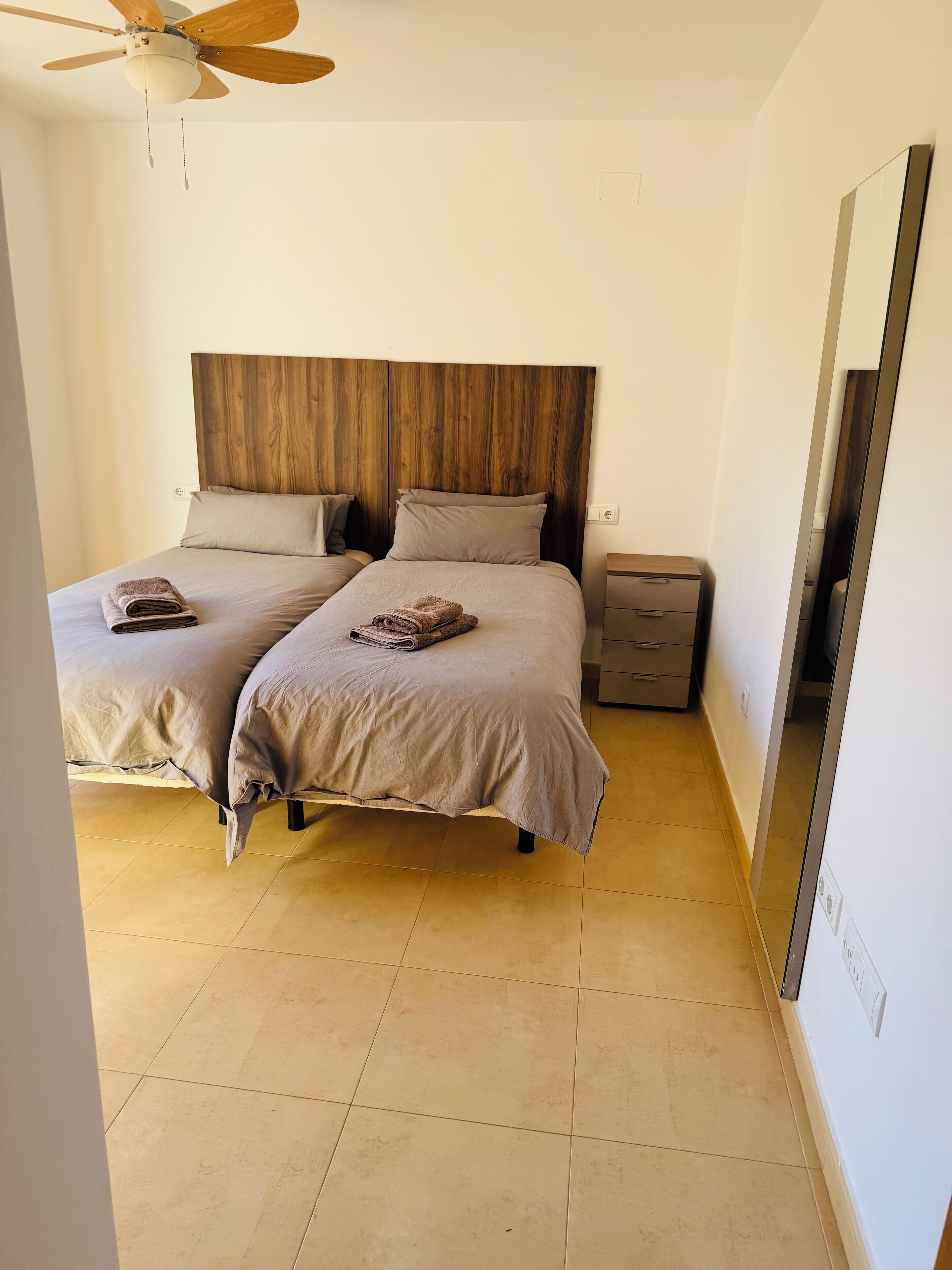 A bedroom with two beds, wooden headboard, bedside tables, and a ceiling fan.