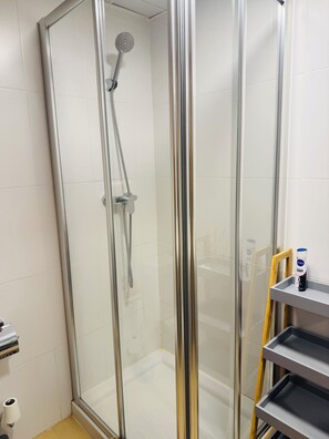 Une douche fermée par des parois en verre, dotée d’un pommeau de douche à main, d’un porte-rouleau de papier toilette et d’une étagère sur laquelle se trouve une bouteille.