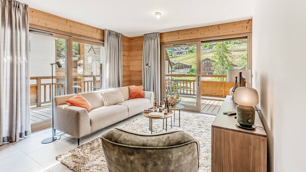 Un salon moderne avec un canapé, un pouf et une table basse, de grandes fenêtres avec des rideaux, et une vue sur une terrasse et des immeubles.