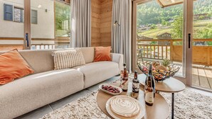 Un salon moderne avec un canapé, une table basse sur laquelle sont posées des bouteilles et une assiette de nourriture, et de grandes fenêtres offrant une vue sur un paysage verdoyant.