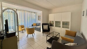 A living room with a sofa, armchairs, a coffee table, and a TV. There is a sliding door with blinds, a cabinet with glass doors, and a framed picture on the wall.