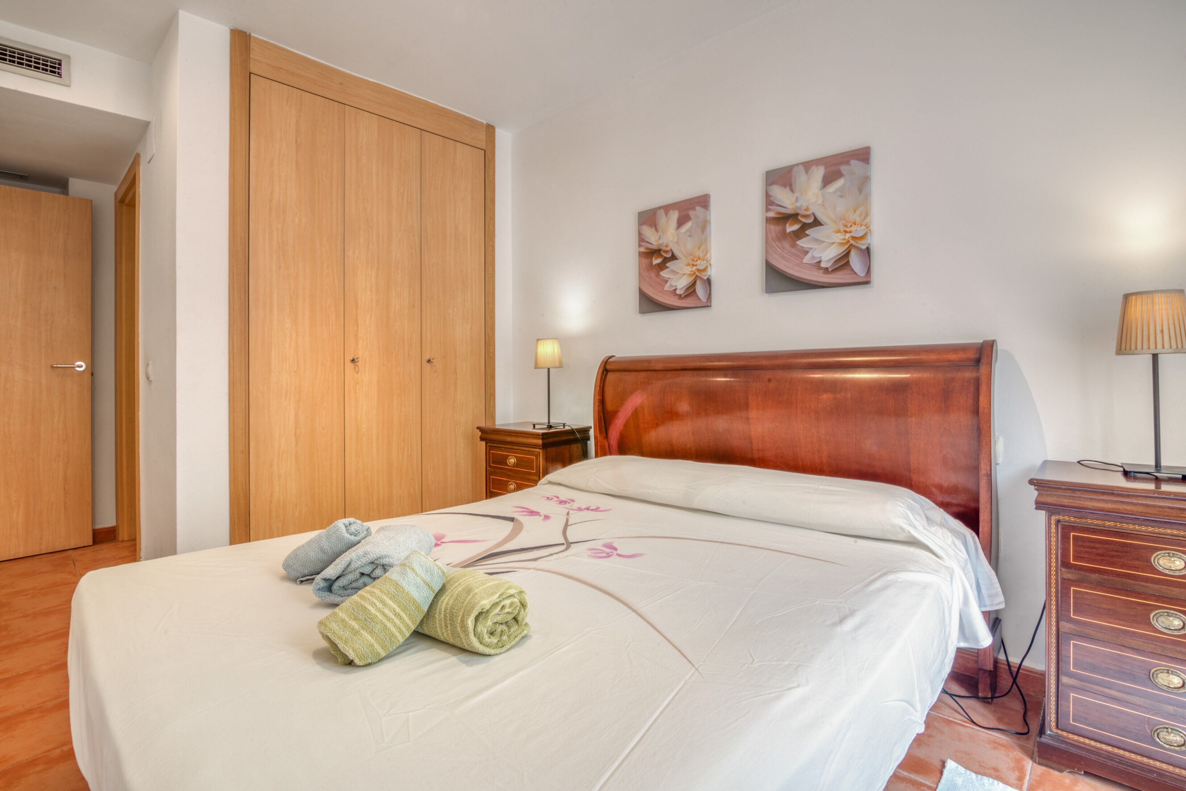 Habitación de hotel con un armario de madera, una cama con colcha estampada floral, dos cuadros de flores en la pared y una mesita de noche con lámpara.