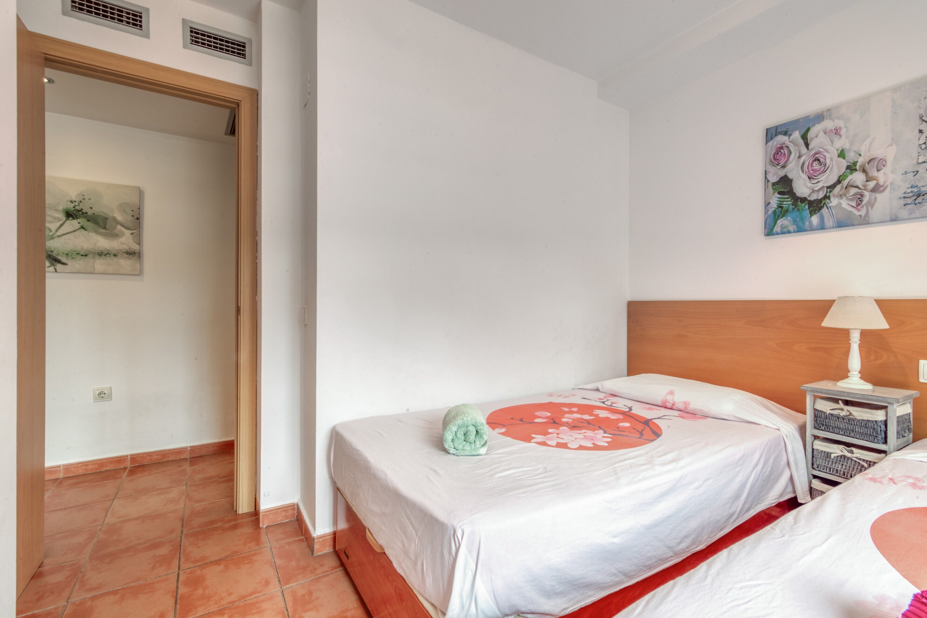 Une chambre avec deux lits, une tête de lit en bois, une table de chevet avec une lampe et un tableau floral accroché au mur.