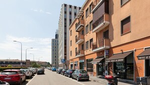 A street with parked cars, a building with a bakery, and a tall apartment building in the background.