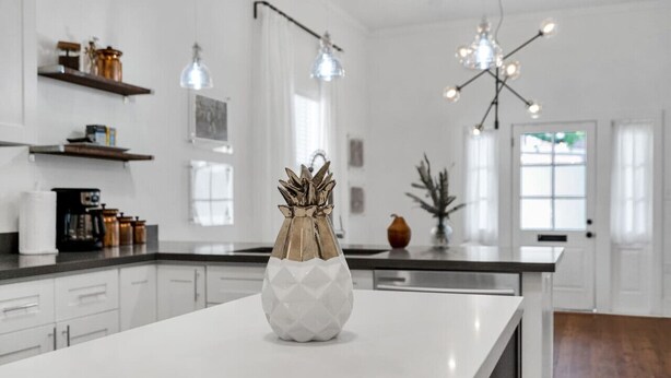 Une cuisine moderne avec des meubles blancs, un îlot central blanc, des luminaires suspendus et des objets décoratifs disposés sur des étagères.