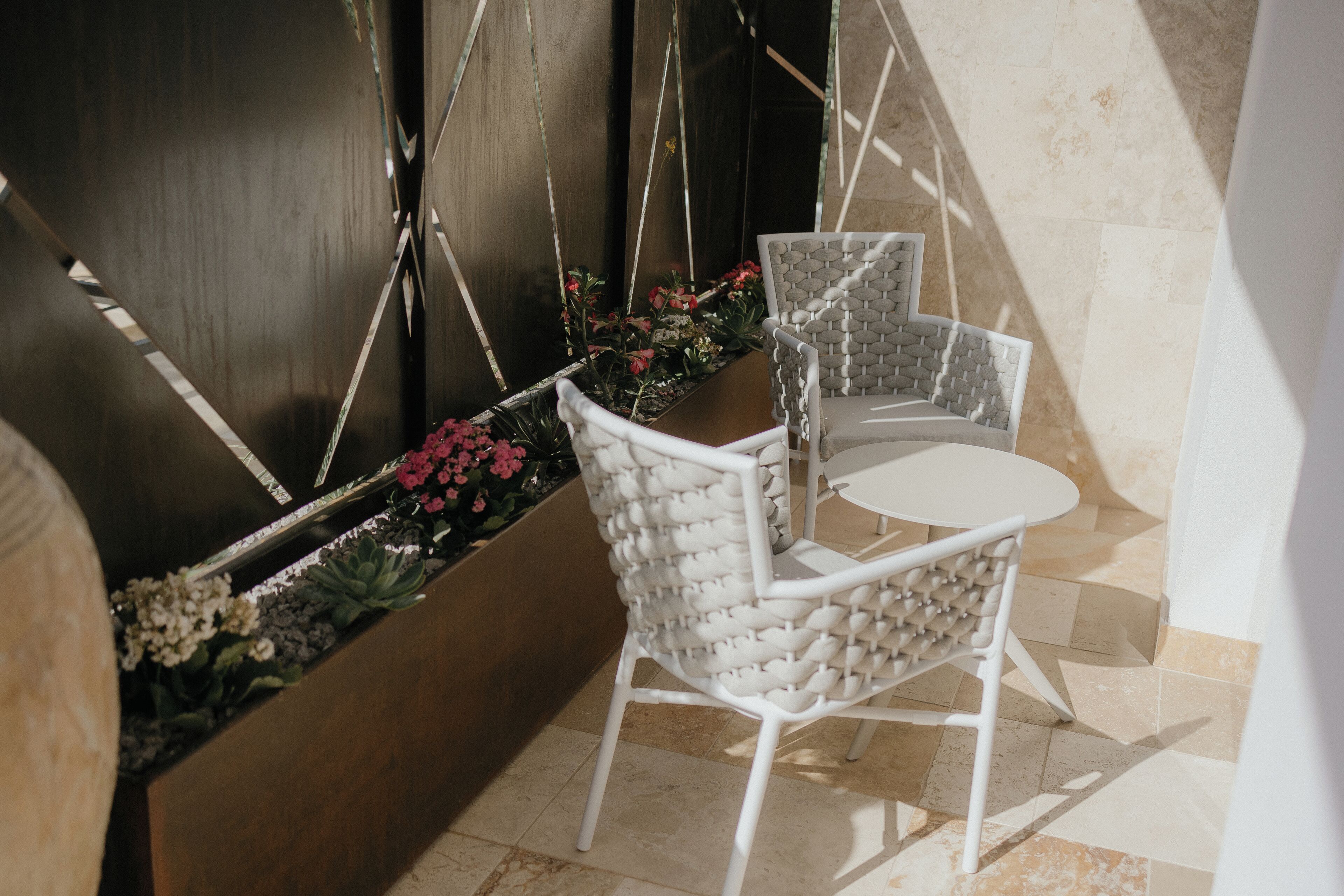 Un petit espace extérieur aménagé avec deux chaises blanches et une table, entouré d’un jardinier fleuri et d’un mur en bois foncé.
