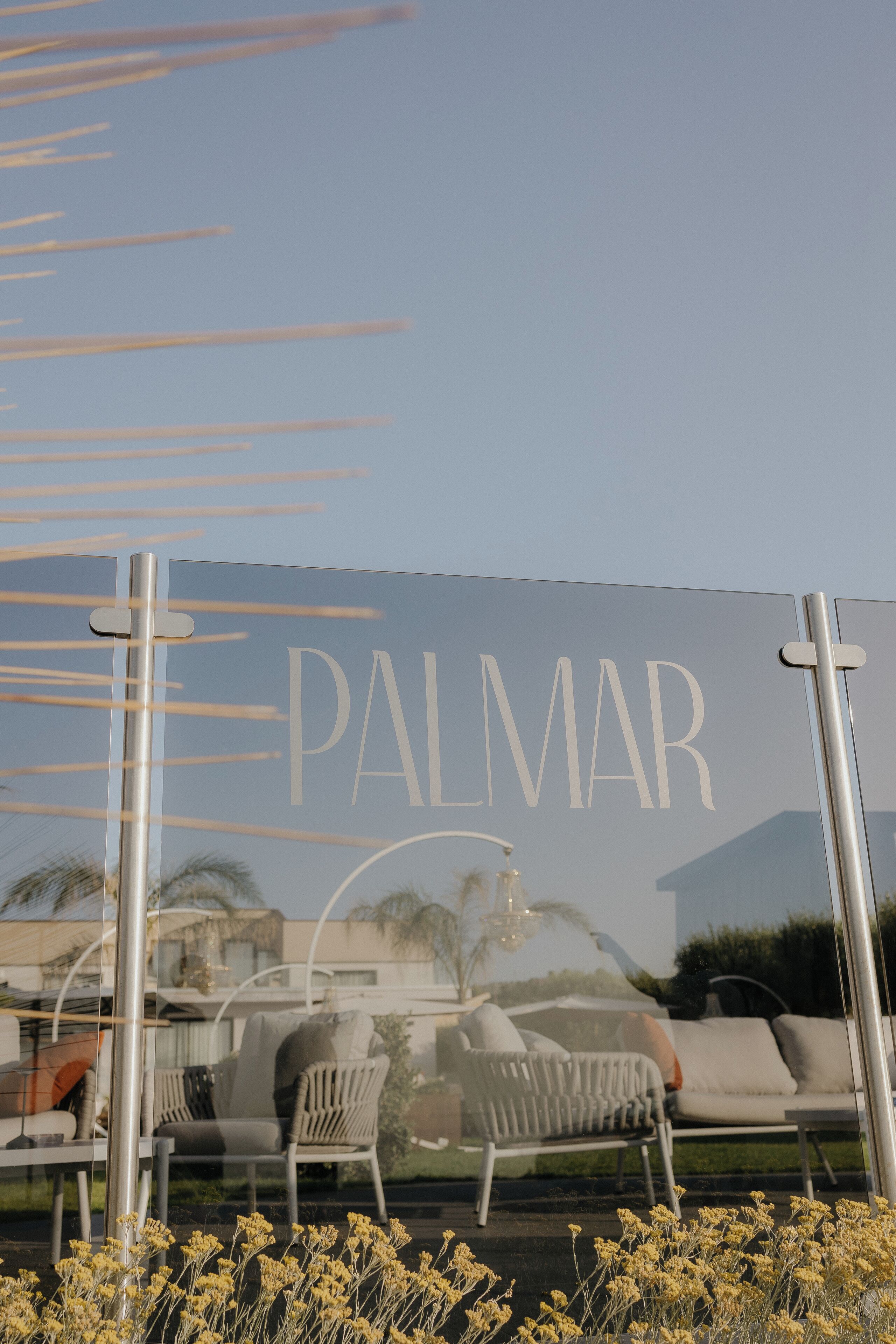 A sign with the word "PALMAR" and a reflection of outdoor furniture and plants.