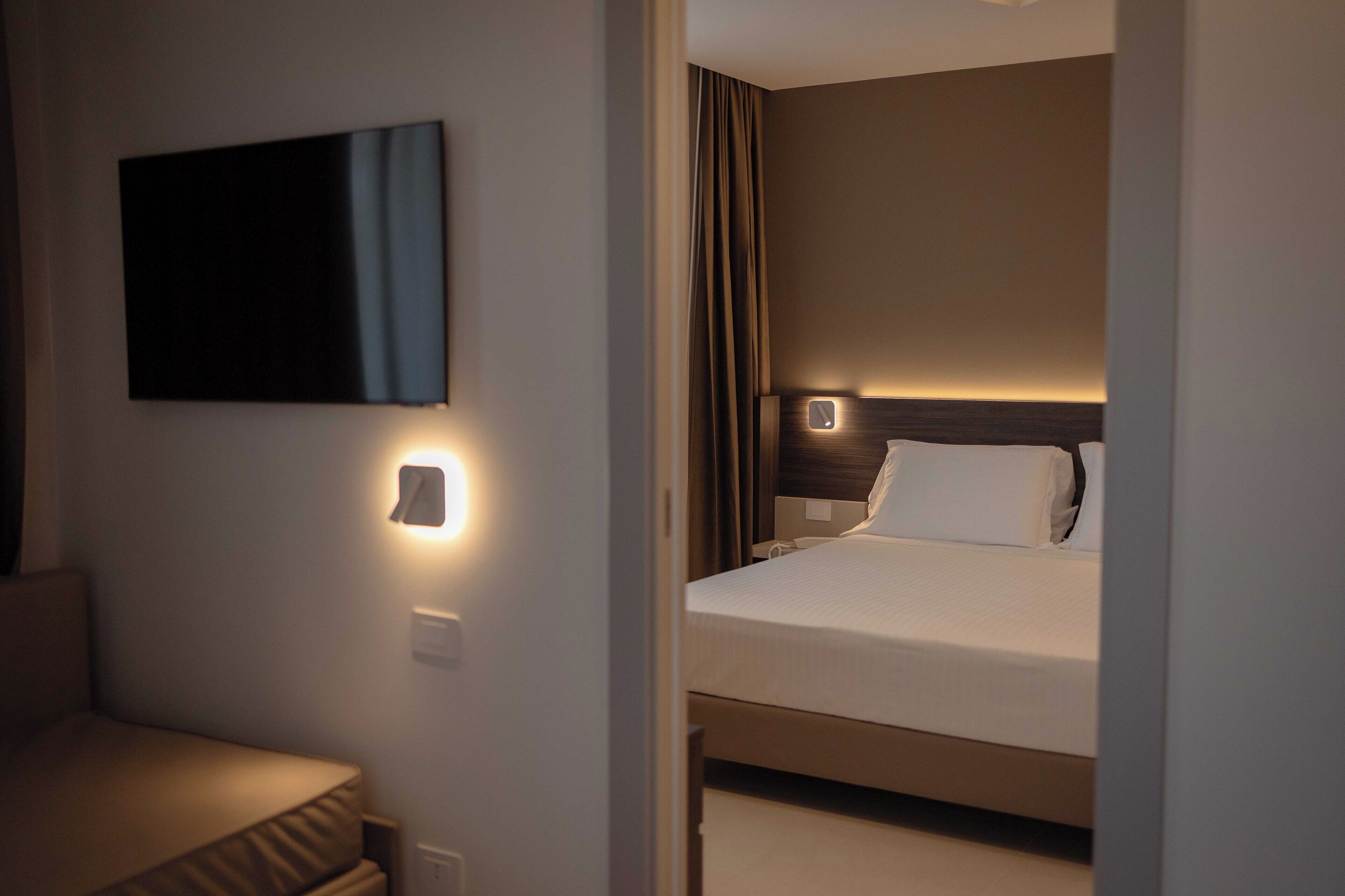 A hotel room with a bed, a bedside table, a wall-mounted TV, and a wall-mounted light.