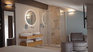 Une chambre d’hôtel moderne dotée d’un grand lit, d’une coiffeuse avec un miroir rond, d  ’un espace douche et d’un coin salon comprenant une chaise et un pouf.