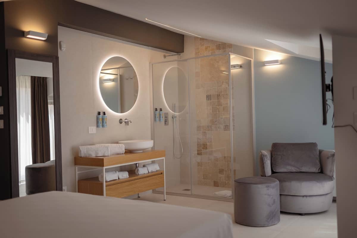 A modern hotel room with a large bed, a vanity with a round mirror, a shower area, and a seating area with a chair and ottoman.