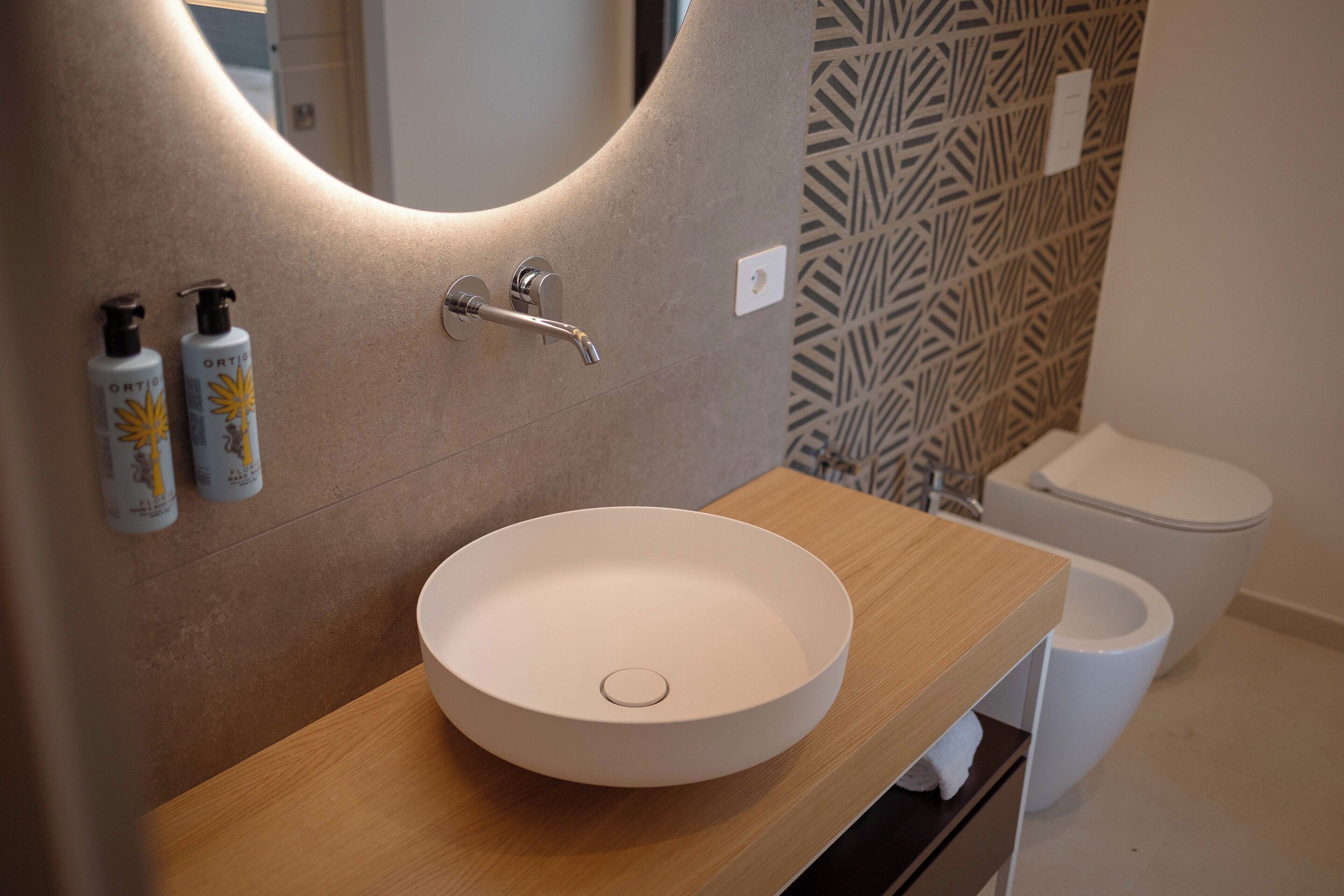A bathroom with a round sink, two bottles of lotion, a faucet, and a toilet.