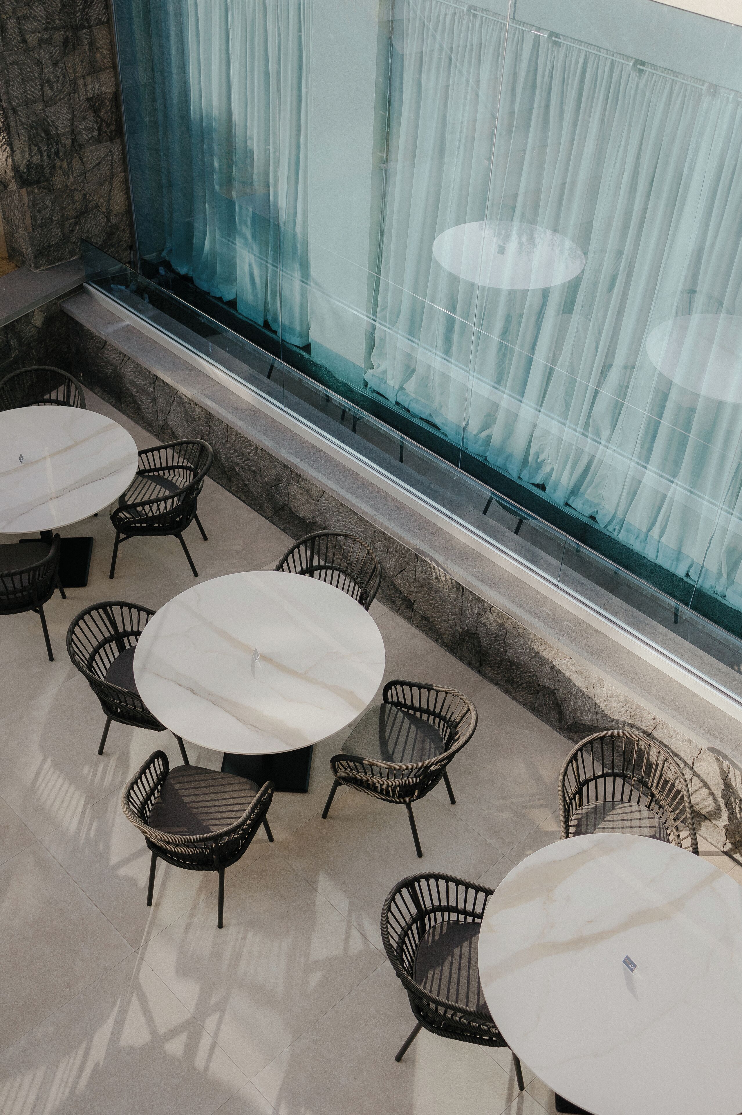 Una terraza con mesas redondas y sillas de mimbre, un muro de piedra y vistas a través de cortinas de tela ligera.