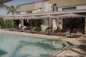 Zona junto a la piscina con sillones de descanso y sombrillas, una piscina cristalina y un edificio moderno al fondo.