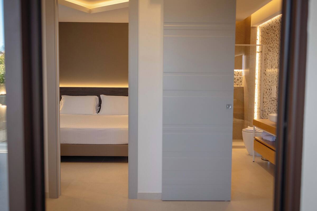 A modern bedroom with a bed, a nightstand, and a bathroom with a toilet and sink.