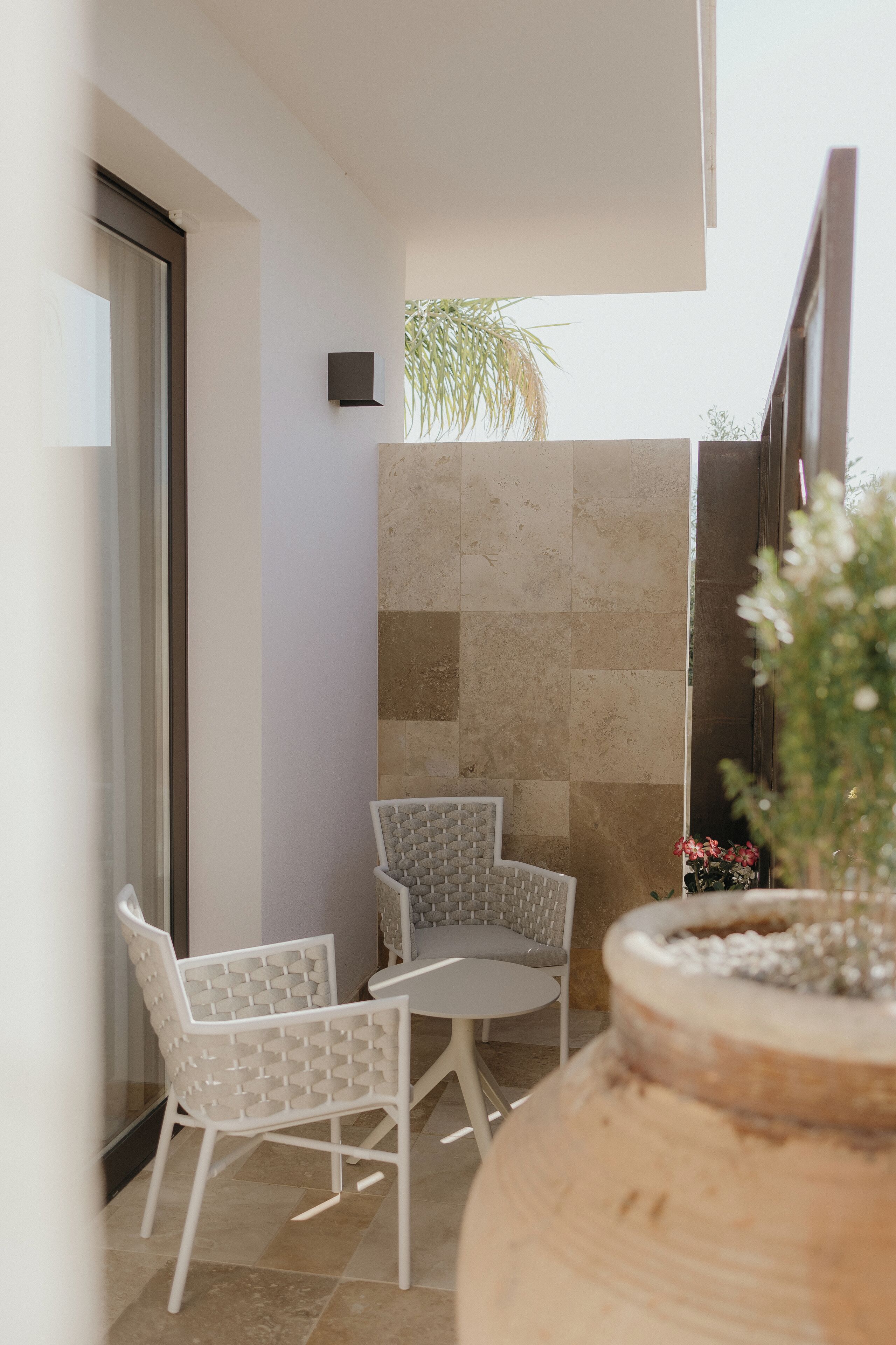 Un petit espace extérieur aménagé avec deux chaises blanches et une table ronde, un pot de fleurs et un mur recouvert de carreaux.