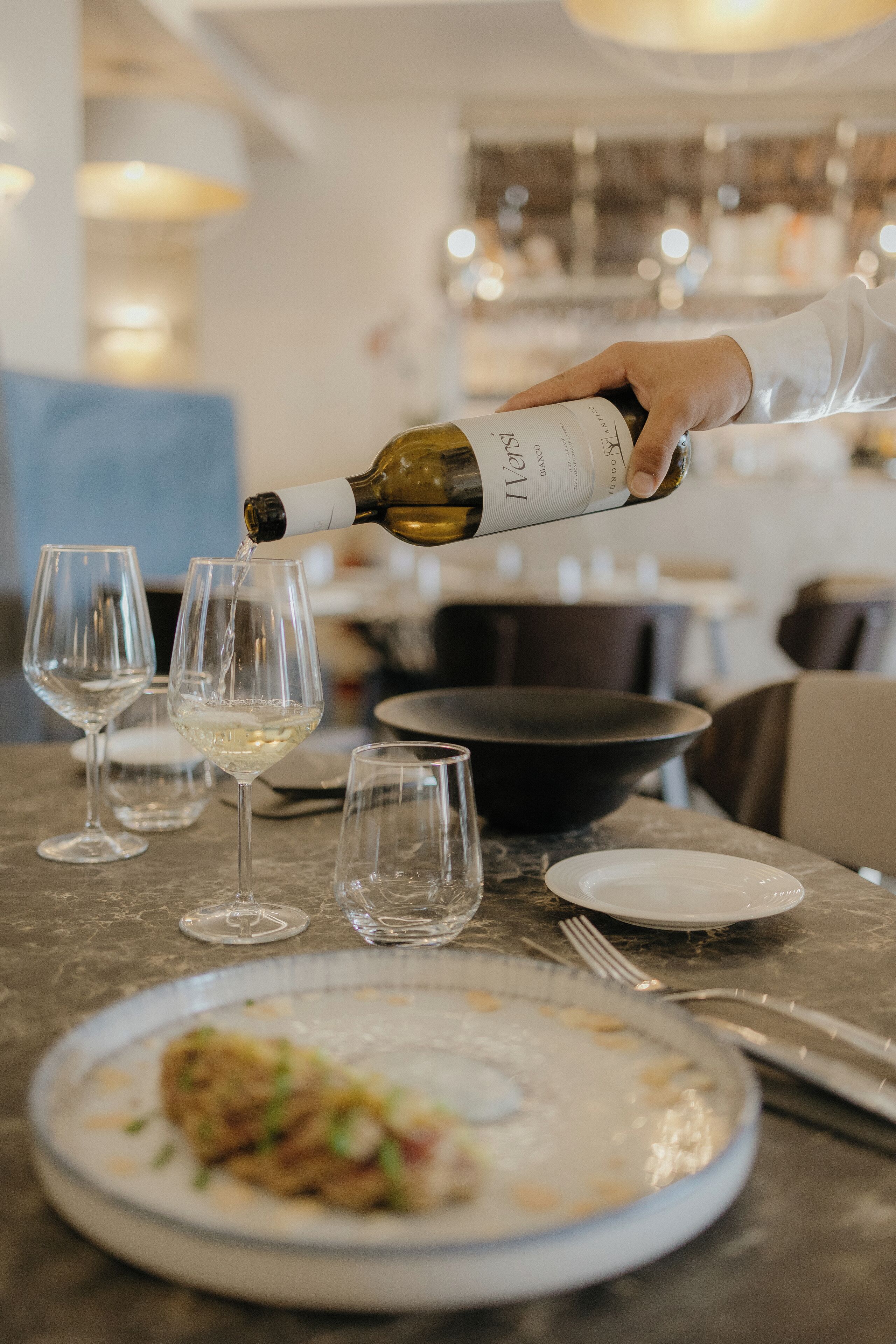 A dining table with wine glasses, a plate of food, and a bottle of wine being poured.