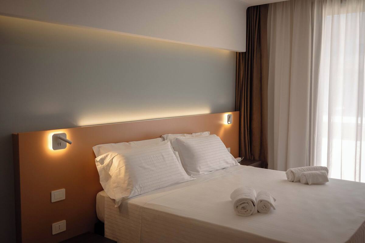 A neatly made bed with white linens and rolled towels, a wall-mounted light, and a window with curtains.