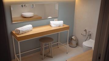 A modern bathroom with a wooden vanity, a round sink, a mirror, a toilet, and a trash can.