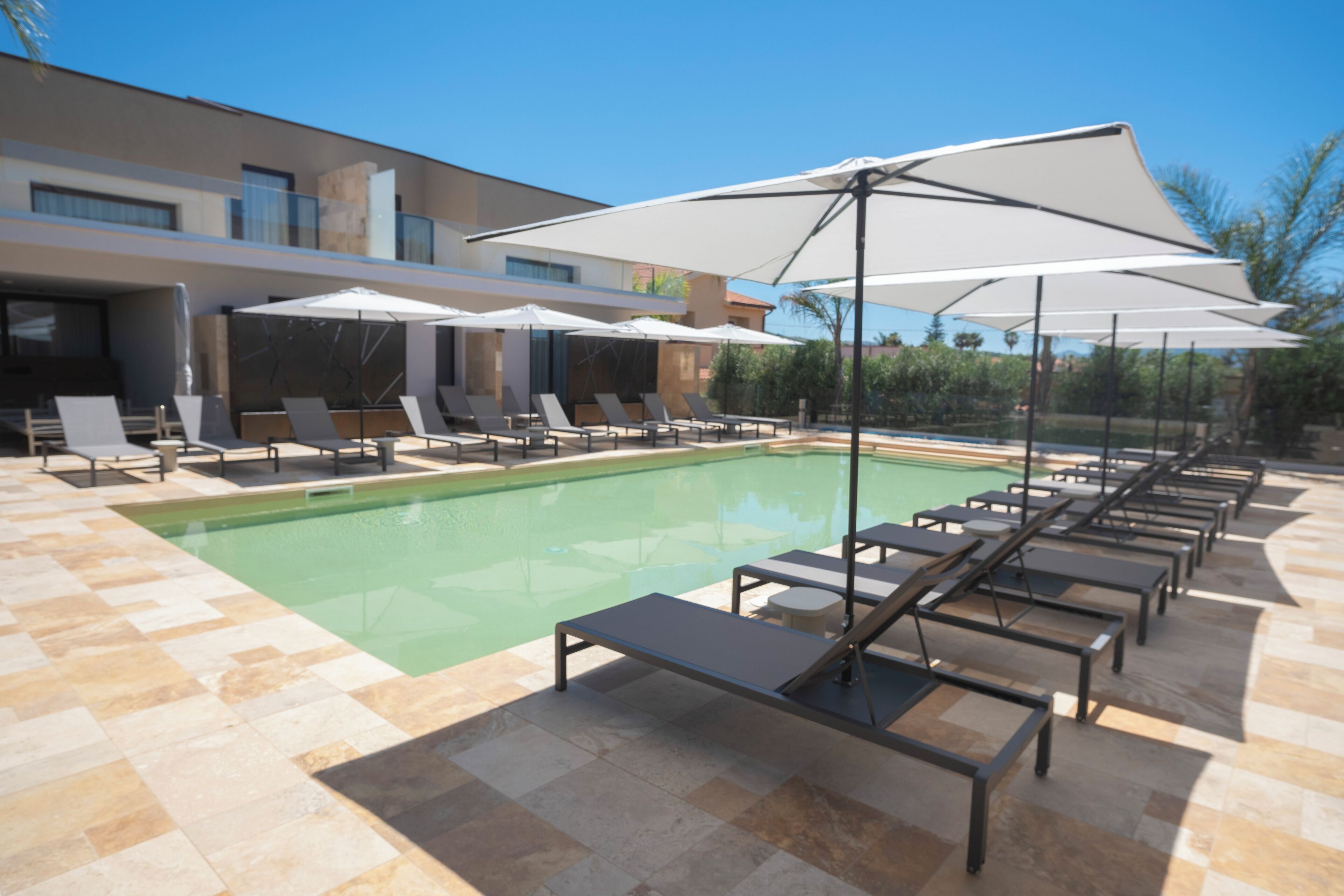 Un espace piscine extérieur moderne, doté de chaises longues et de parasols, entouré d’une terrasse carrelée et d’un bâtiment aux grandes fenêtres.