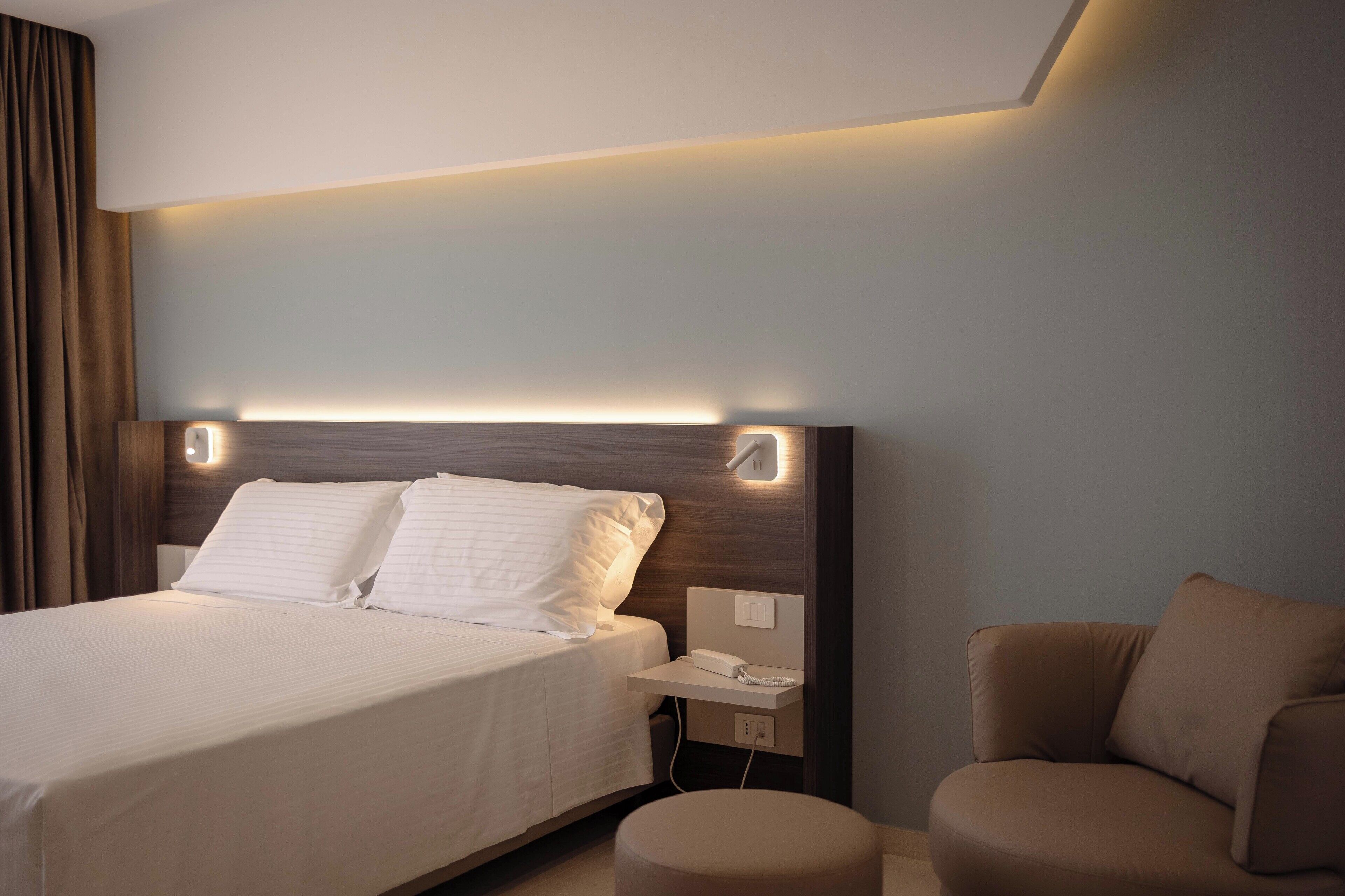 Moderni hotellihuone, jossa on siististi pedattu sänky, yöpöytä, jossa on puhelin, ja ruskea nojatuoli.