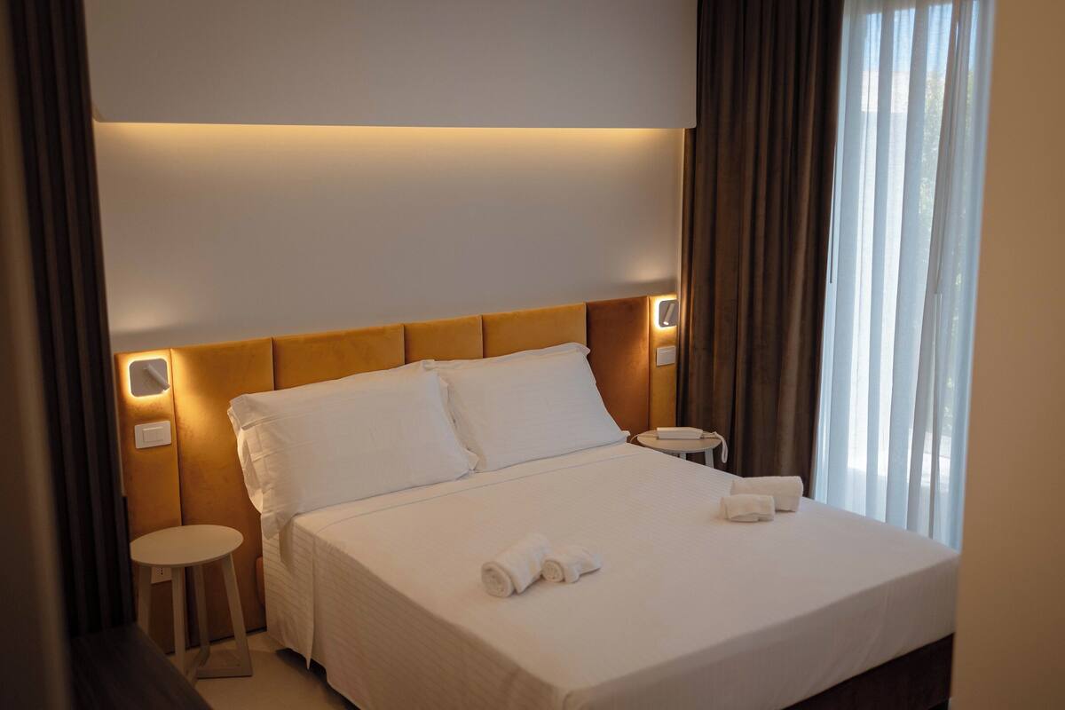 A neatly made bed with white linens and towels, a round bedside table, and a wall-mounted light fixture.
