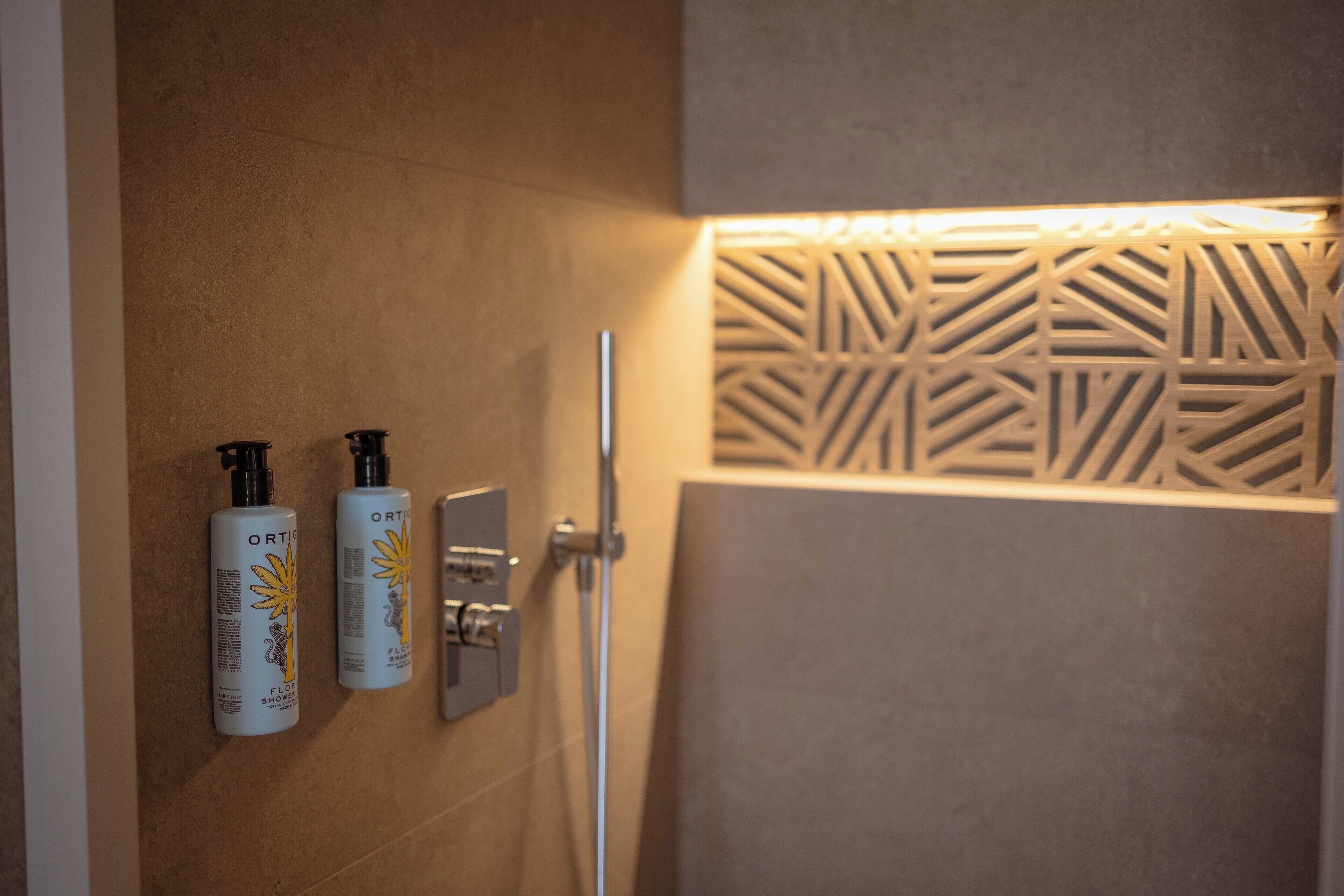 Casa de banho com duas embalagens de gel de banho Ortex, um chuveiro e um painel decorativo na parede.