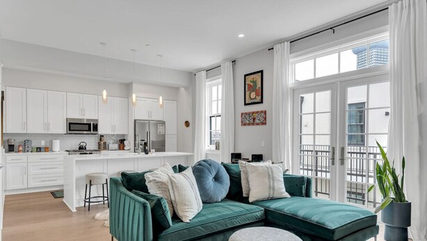 A modern apartment with a green sofa, a kitchen with white cabinets, and large windows.