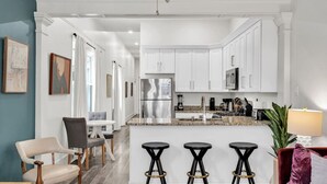 A modern kitchen with white cabinets, a stainless steel refrigerator, and a granite countertop. There are three black bar stools and a red sofa in the living area.