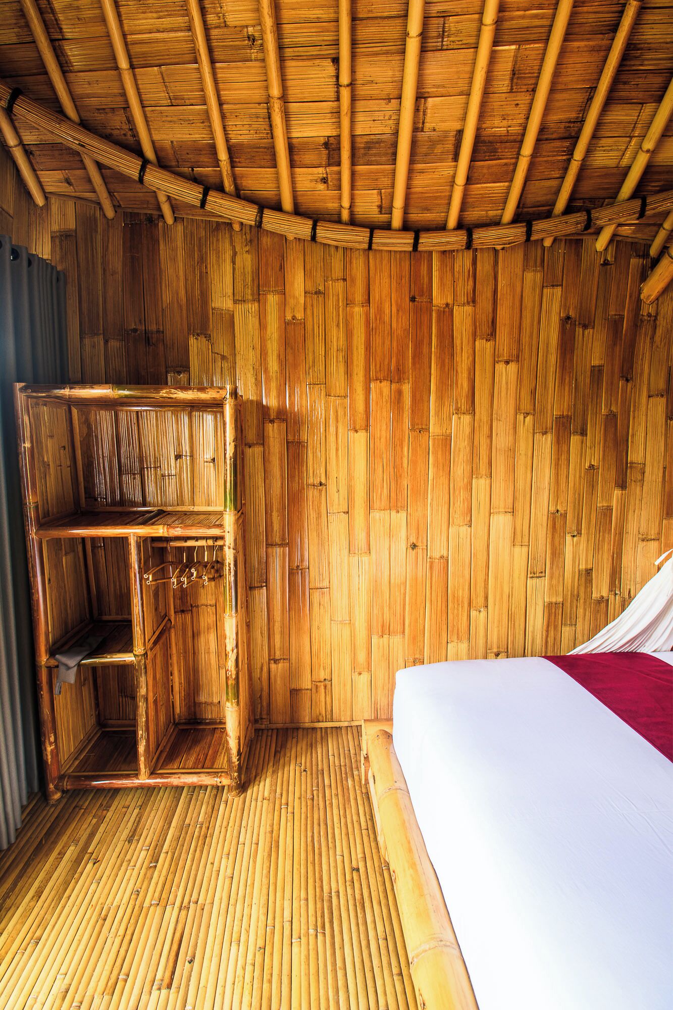 A room with a bamboo wall and floor, a bed with a white and red bedspread, and a wooden wardrobe with a mirror.