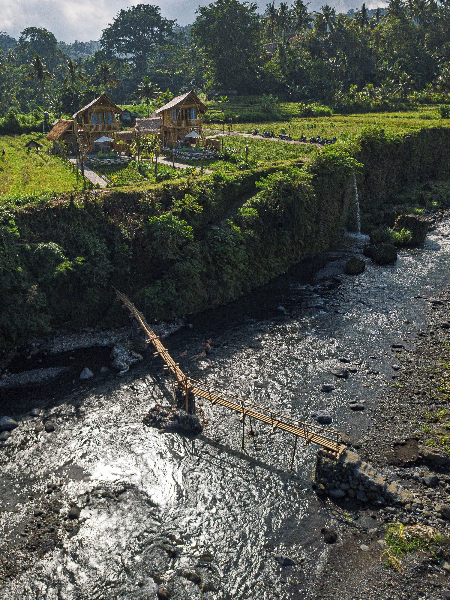 A rustic wooden bridge over a rocky stream, leading to a wooden house with a balcony, surrounded by lush greenery and a waterfall.