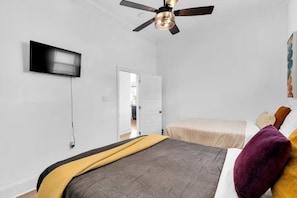 Un dormitorio con cama, ventilador de techo, televisor montado en la pared y una puerta que da a otra habitación.