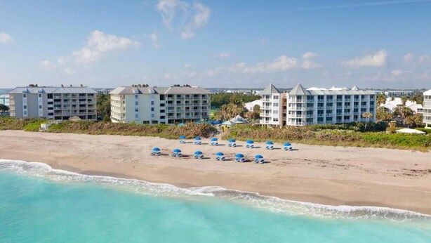 Et hotel ved stranden med flere bygninger, en sandstrand og flere blå parasoller.