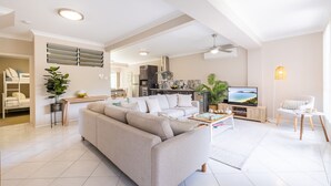 Un salon spacieux avec un canapé, une table basse et un téléviseur. On aperçoit une cuisine au fond et un balcon avec vue.