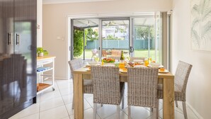 Un coin repas avec une table dressée pour quatre personnes, entouré de chaises en osier, une porte-fenêtre coulissante donnant sur un jardin, et un espace cuisine avec un réfrigérateur et des placards.