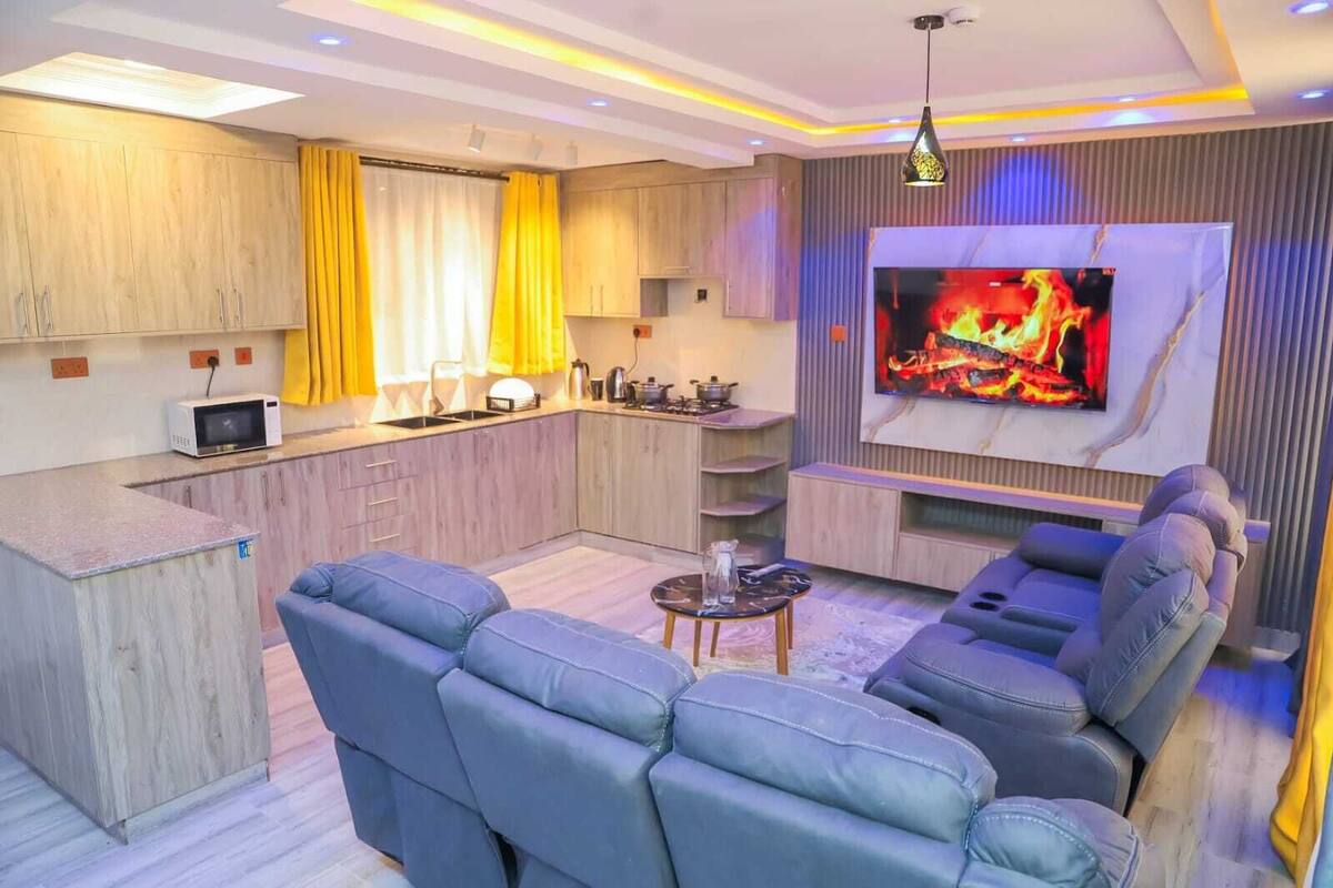 A modern living room with a large TV, a sofa, a coffee table, and a kitchen area with wooden cabinets and yellow curtains.