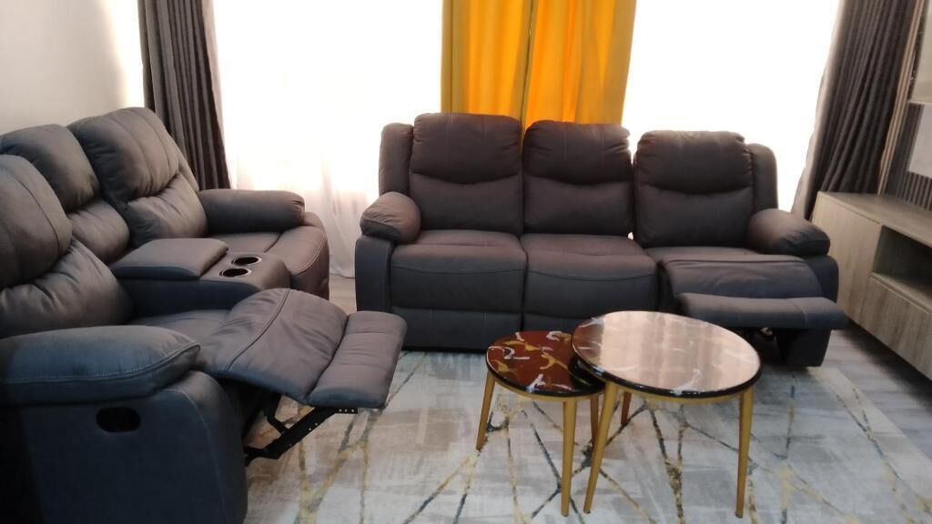 A living room with a dark-colored sofa, a recliner, and a small round table with a patterned cushion.