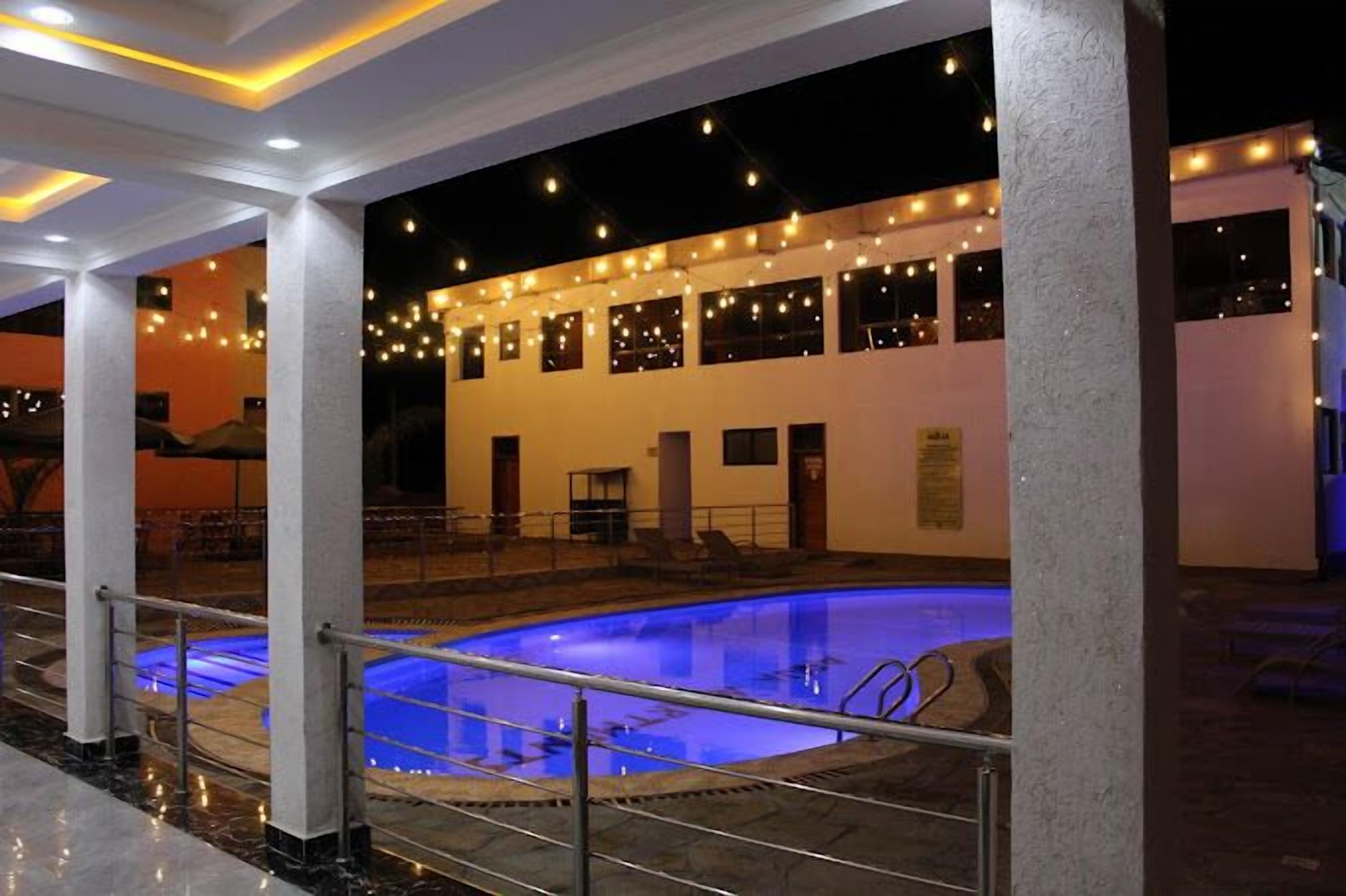 Una piscina interior con un tono azul, rodeada por un área techada con columnas y luces colgantes.