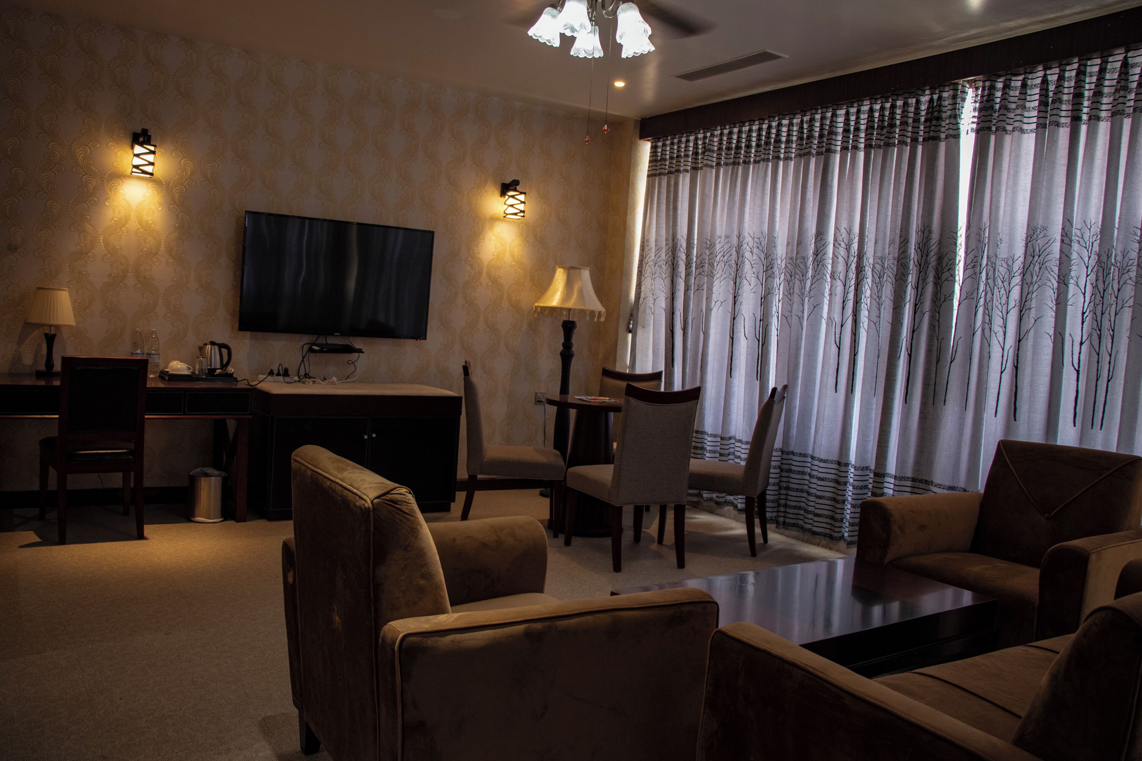 A hotel room with a flat-screen TV, a desk with a lamp, a dining table with chairs, and a sofa.