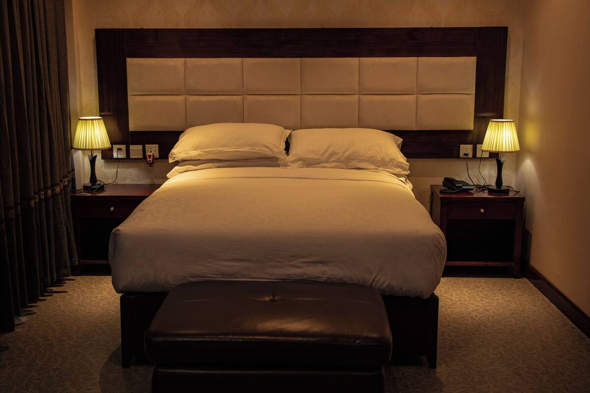 A hotel room with a large bed, two bedside lamps, a nightstand, and a bench.
