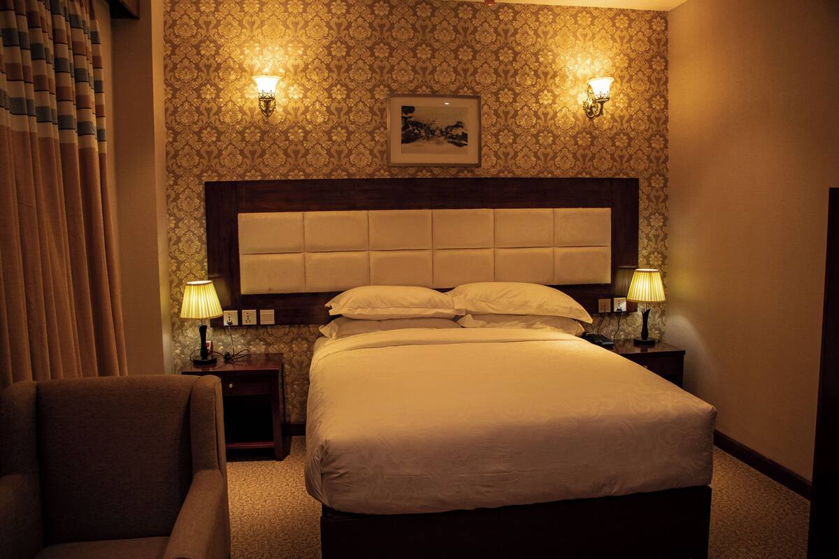 A hotel room with a large bed, two bedside lamps, a headboard, a framed picture on the wall, and a chair.