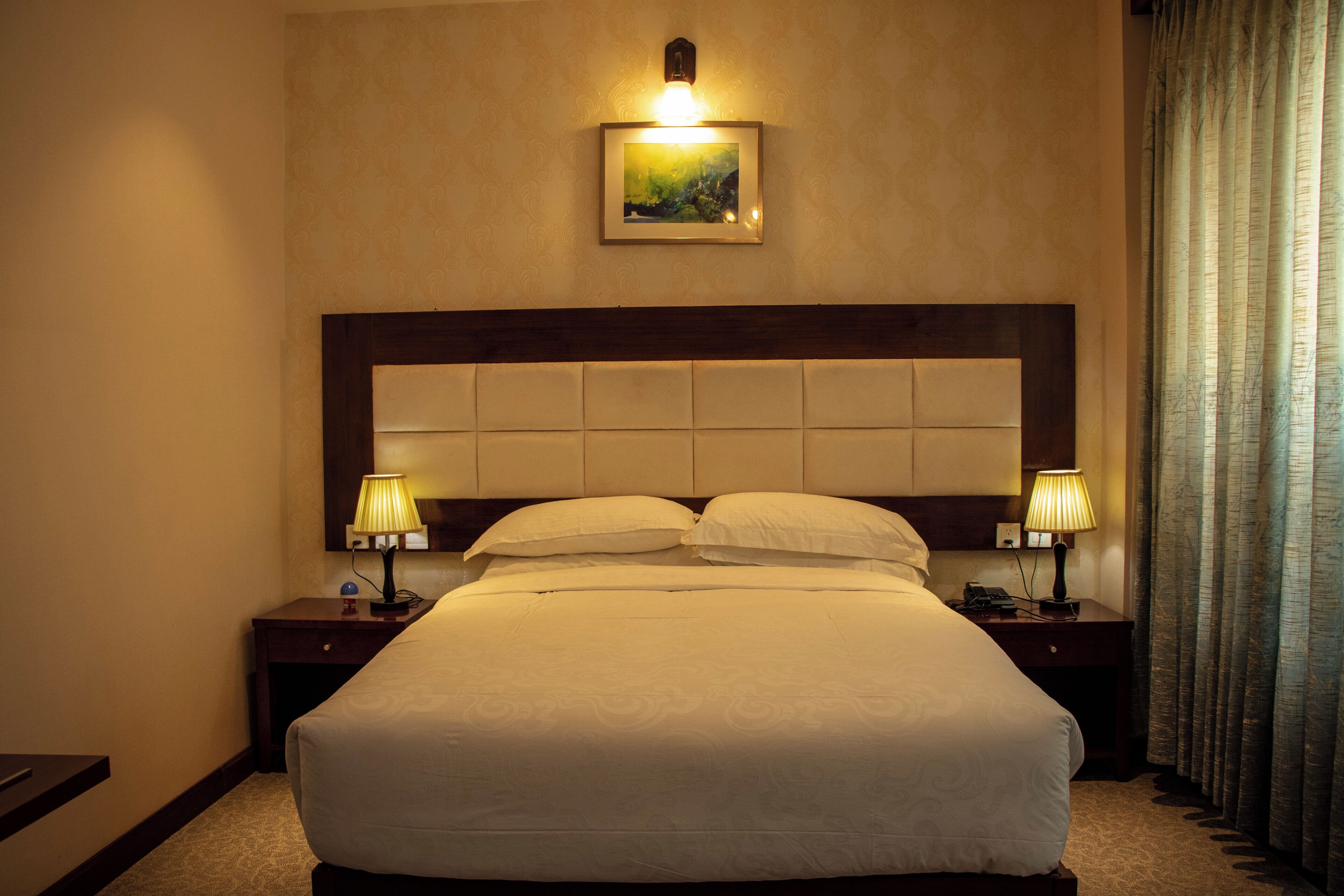 Quarto de hotel com uma cama grande, duas mesas de cabeceira com luminárias, cabeceira e um quadro na parede.