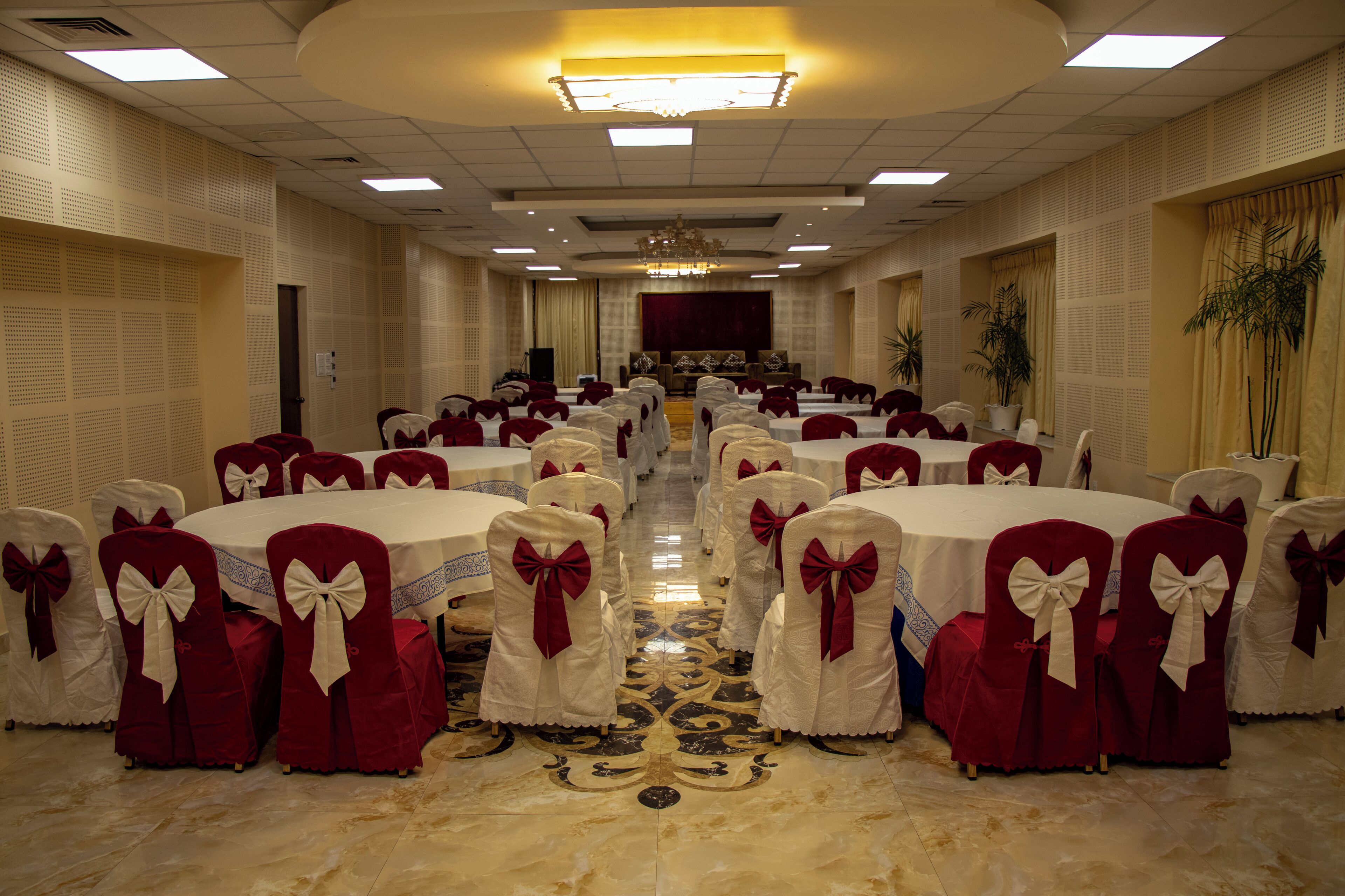 A banquet hall with round tables, red and white chairs with bows, a chandelier, and a stage at the front.