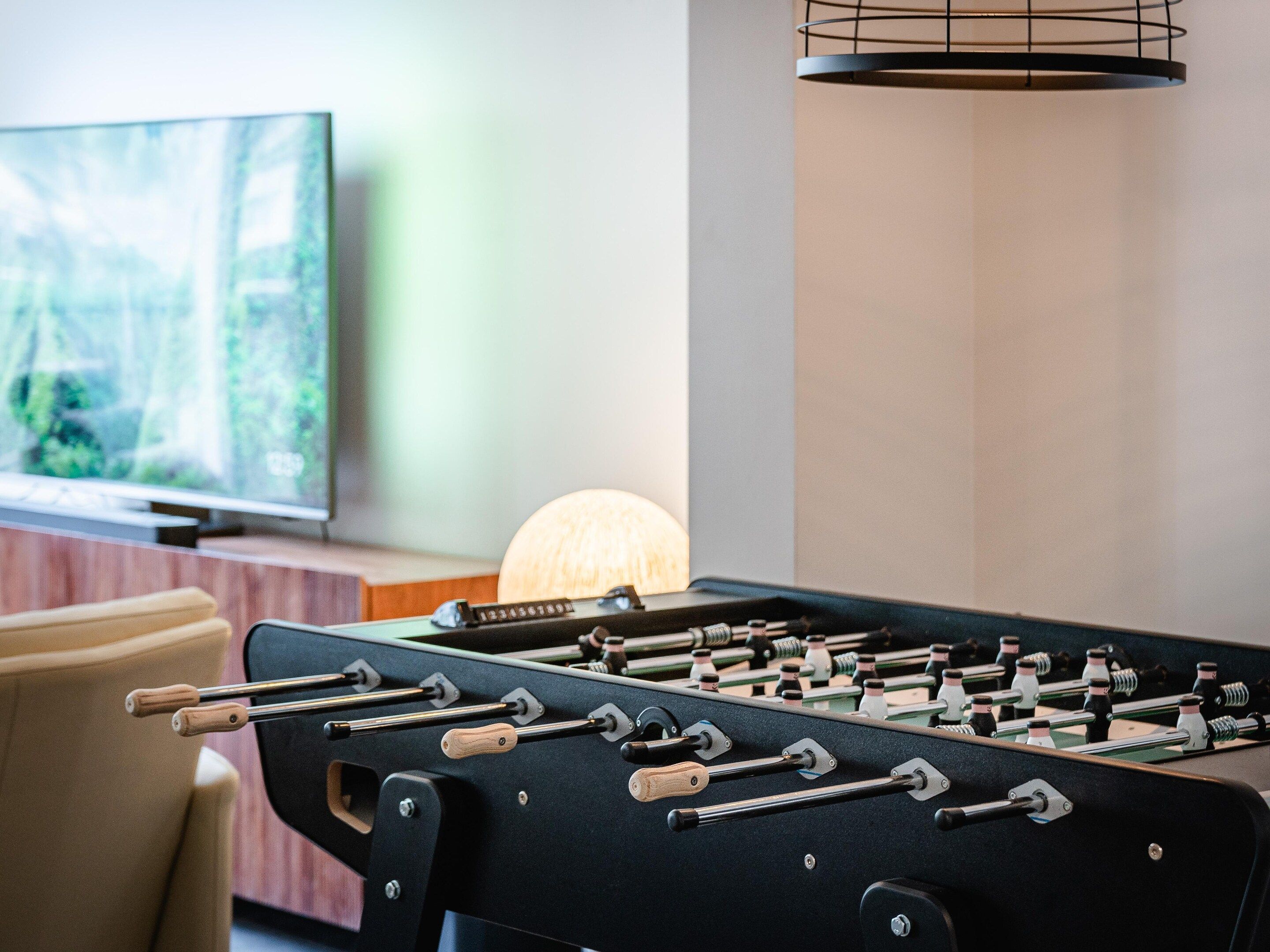 A foosball table with a modern design, a television in the background, and a lamp on a table.