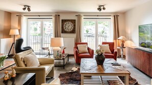 A modern living room with leather sofas, a coffee table, and a TV. There are two windows with curtains, a clock on the wall, and a radiator. - Vacation Home in Bouillon by Semois River (Bouillon)
