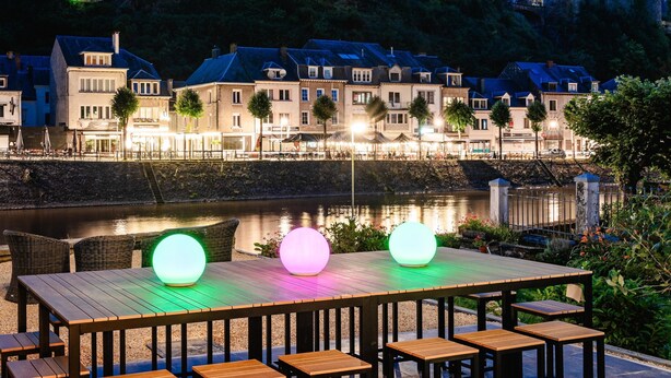 A riverside view of a historic town with a castle on a hill, wooden tables with glowing orbs, and a calm river. - Vacation Home in Bouillon by Semois River (Bouillon)