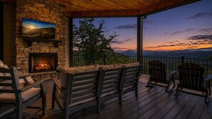 A wooden deck with a stone fireplace, outdoor seating, and a view of the mountains at dusk.