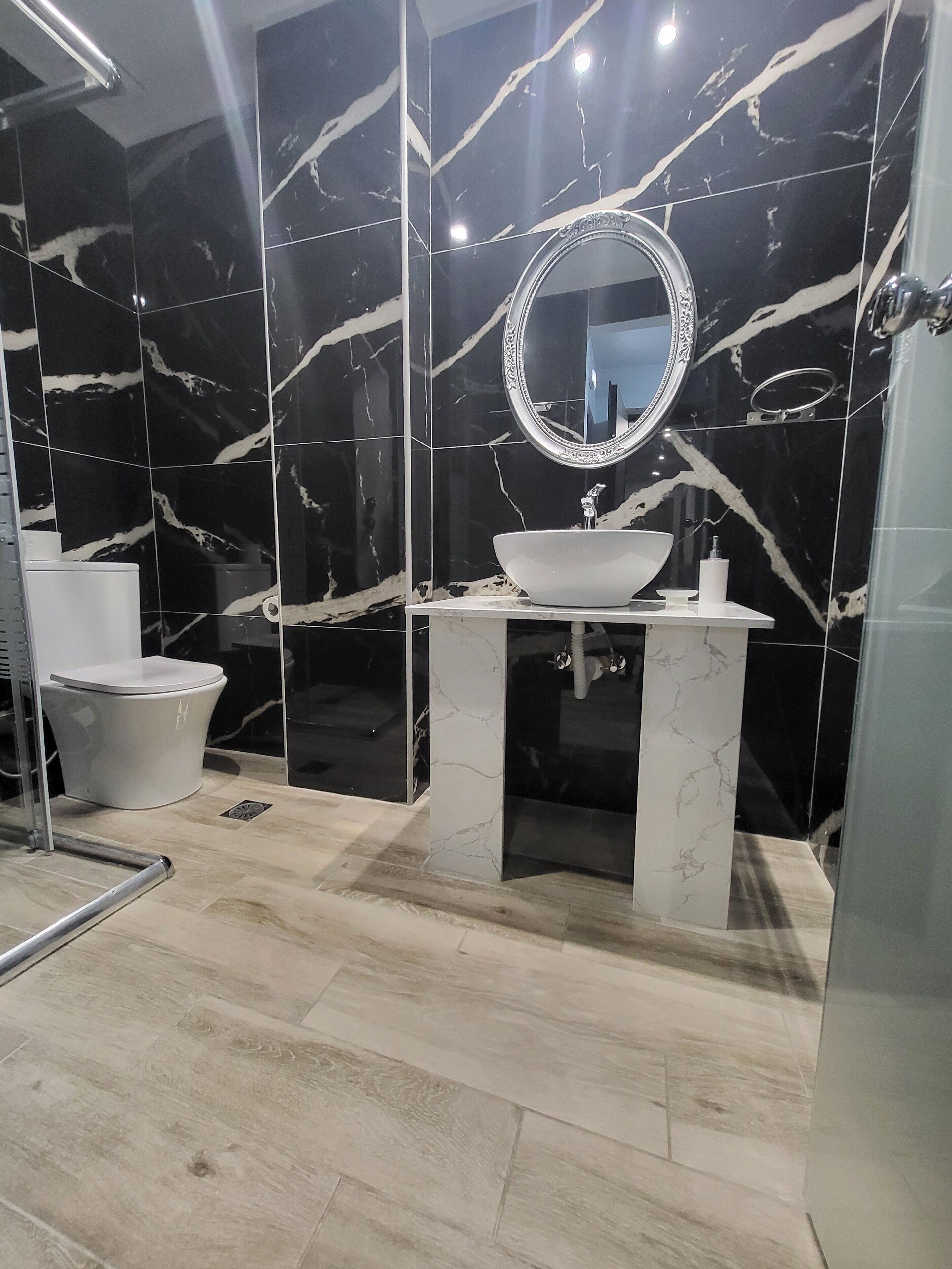 Une salle de bain moderne avec un pare-baignoire en marbre, un miroir rond, un lavabo blanc et des toilettes.