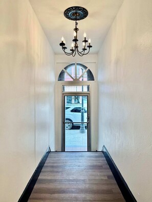 A hallway with a chandelier, a door with a window, and a view of a parked car outside.
